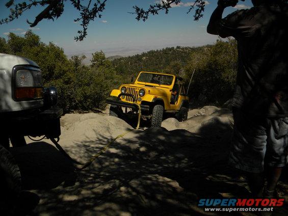 Yellow Jeep Wrangler on rocks, white truck in foreground. Off-road scene