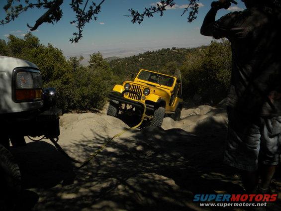 Yellow Jeep Wrangler on rocky trail, white truck in foreground