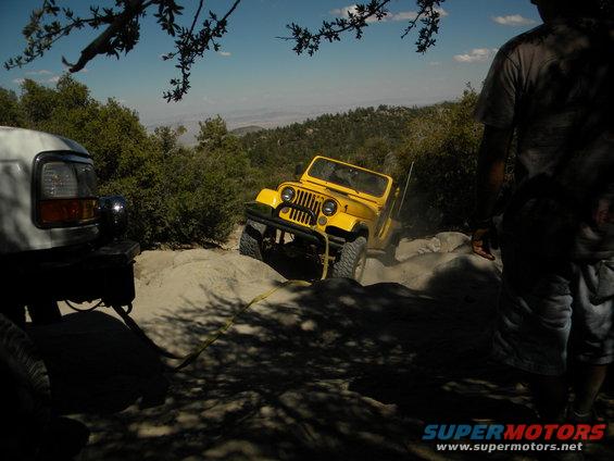 Yellow Jeep Wrangler navigating rocky terrain, white truck in foreground