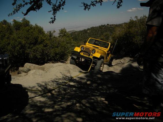 Yellow Jeep Wrangler on rocky terrain, front bumper visible, with a tow rope attached