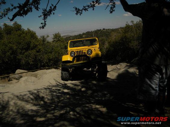 Yellow Jeep CJ driving on a rocky trail with trees and a person in the foreground