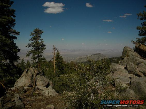 Mountain landscape with trees, rocks, and a distant valley under a blue sky with clouds. Watermark reads www.supermotors.net