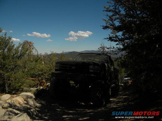1992 Ford Bronco driving on a rocky dirt road with trees and mountains in the background. Another vehicle is visible ahead