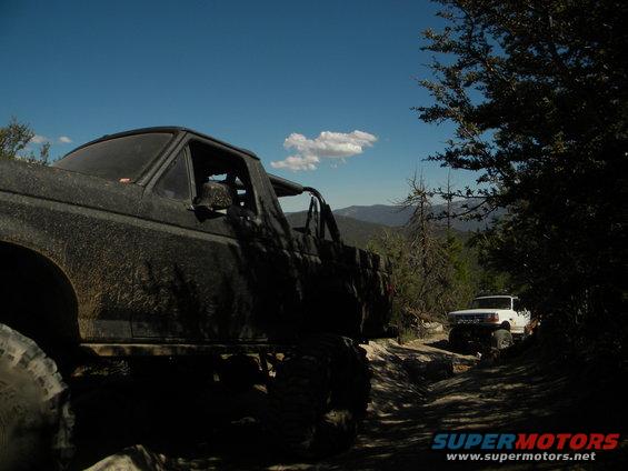 Muddy off-road truck in foreground, white SUV on trail behind it, trees and mountains in background