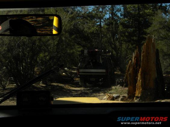View from inside a vehicle looking at another SUV driving on a dirt road in a forest. The SUV has a roof rack