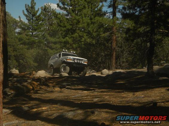 1992 Ford Bronco driving over rocks on a wooded trail. It has aftermarket lights on the roof and front