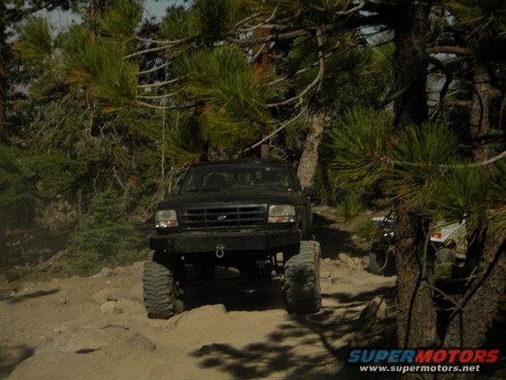 Black SUV with large tires driving on a rocky trail, trees around. Another vehicle is partially visible to the right