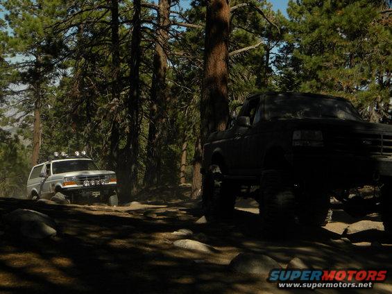 White 1992 Ford Bronco with roof lights drives on a dirt path in a forest, with a dark truck in the foreground