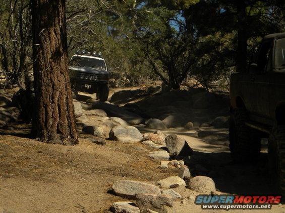 Two off-road vehicles driving on a rocky trail in a wooded area. One vehicle is in the foreground on the right, and another is further up the trail on the left