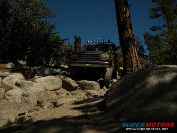 1992 Ford Bronco navigating a boulder-strewn trail with trees and clear blue sky