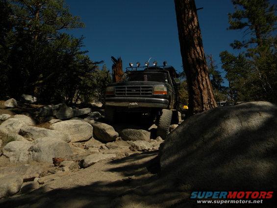 Dark colored 1992 Ford Bronco navigating a rocky trail with trees and a clear blue sky