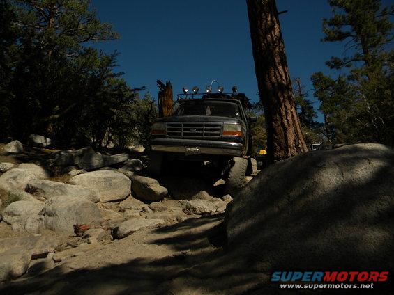 Dark 1992 Ford Bronco navigating a rocky, off-road trail with trees and blue sky