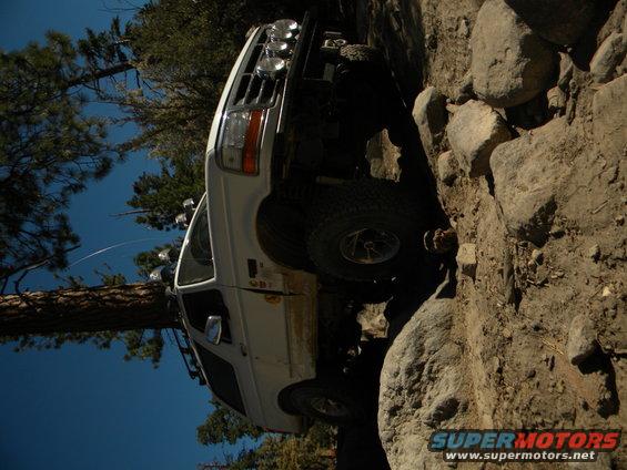 White SUV is climbing a rocky incline with trees and blue sky in the background. It has off-road tires and extra lights on the roof