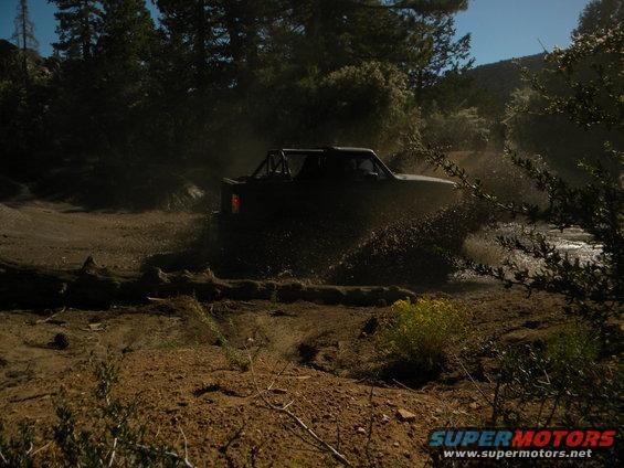 Dark off-road vehicle with roll cage driving through mud and water, kicking up spray