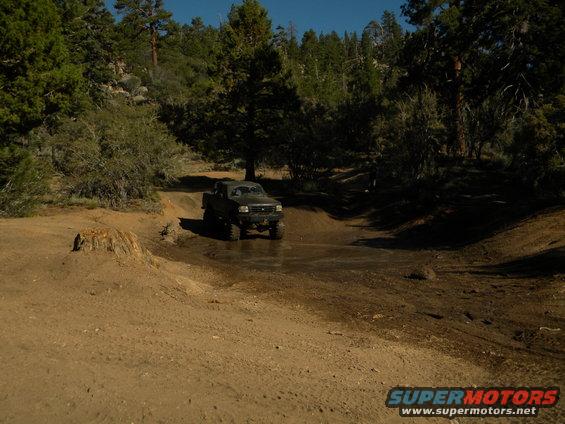 Dark colored pickup truck driving through a muddy puddle on a dirt trail with trees in the background