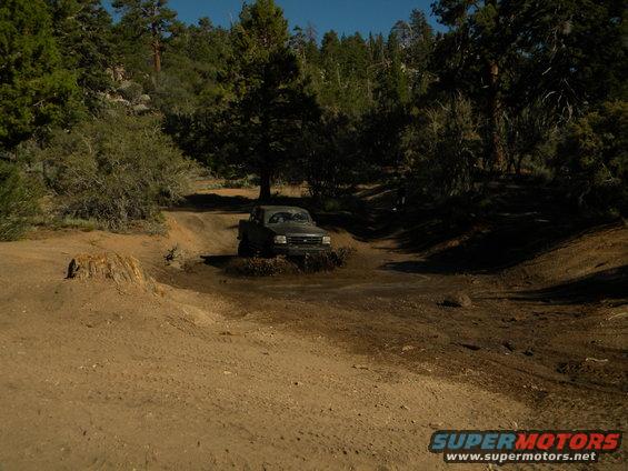 Dark-colored truck driving through mud on a dirt road with trees in the background