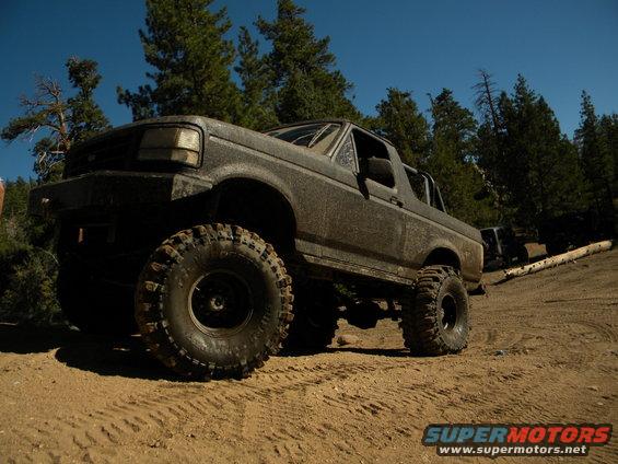 Muddy SUV with large tires on a dirt road, trees in background. Another SUV is visible behind it