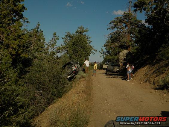 Off-road vehicle overturned on a dirt embankment next to a dirt road with other vehicles and people