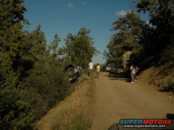 Overturned SUV on a dirt road with people nearby and another vehicle on the road