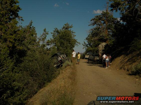 Off-road vehicle on its side in bushes next to a dirt road with people standing nearby