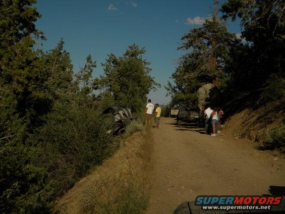 Off-road vehicle tipped into bushes on a dirt road with people standing around