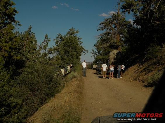 Camouflaged off-road vehicle tilted on a dirt embankment next to a wooded area