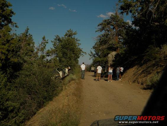 Off-road vehicle partially stuck in bushes on a dirt road with people standing nearby