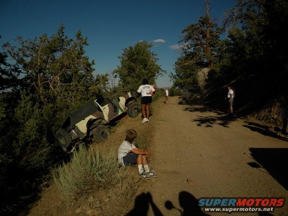 Camouflaged off-road vehicle tilted on a dirt embankment, with people standing on the road