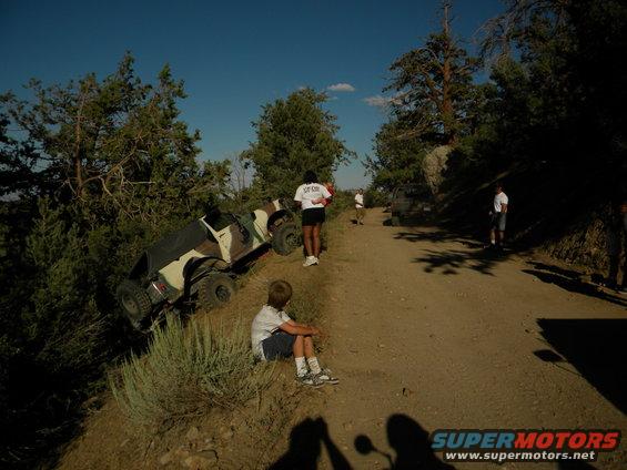 Camouflaged SUV tilted on its side on a dirt road with people nearby