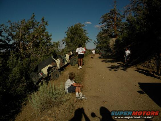 Camouflaged off-road vehicle tilted on a steep dirt road with people nearby