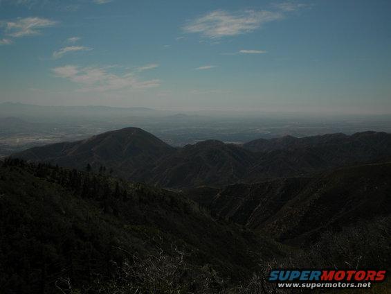 View of hazy mountains and a distant valley under a blue sky with scattered clouds. Supermotors.net logo in the corner