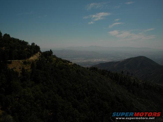 Mountainous landscape with a valley and distant haze under a blue sky with clouds. Watermark at bottom right