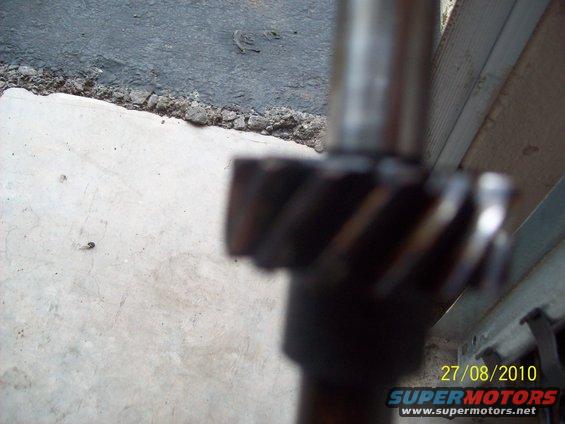 Close-up of a metal gear with helical teeth on a shaft, with concrete and a wooden structure in the background