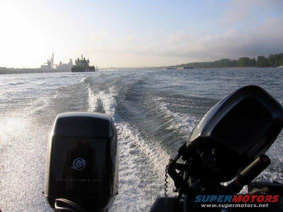 Two black outboard boat motors on choppy water, with a barge and industrial buildings in the distance