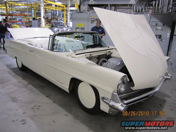 Cream-colored convertible with hood open, showing engine. Interior is white. Man in blue shirt stands nearby