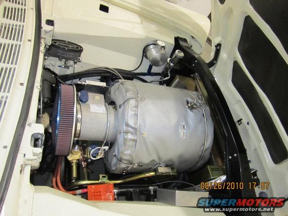 Engine bay with a large, insulated engine component, air filter, and various hoses. White exterior paint visible