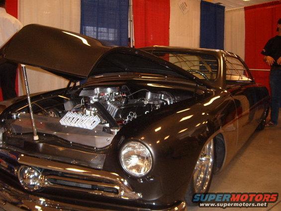 Dark brown hot rod with hood open showing a V8 engine with dual carburetors. Chrome grille and headlights visible