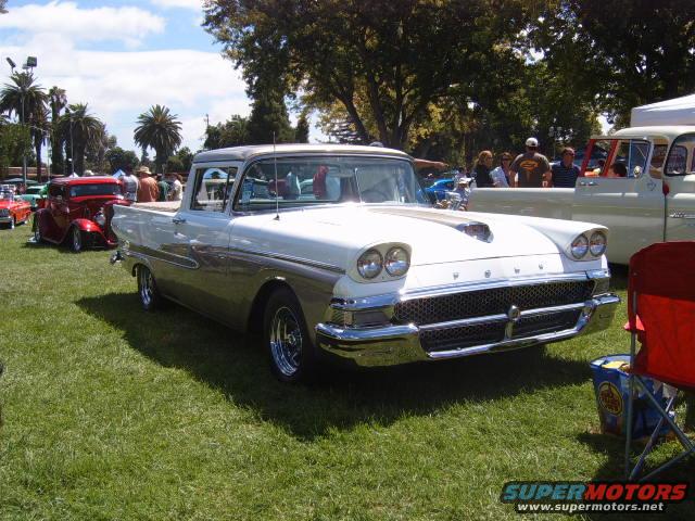 White and tan Ford Ranchero pickup with a hood scoop parked on grass at a car show. Another red hot rod is visible to the left
