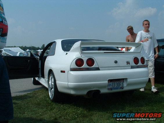 White coupe with a big rear spoiler and dual taillights. Two guys stand nearby