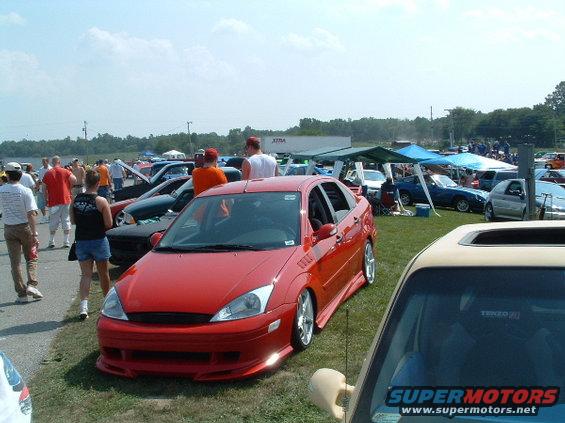 Red sedan with custom body kit parked on grass at an outdoor car show. Several other cars and people are visible in the background