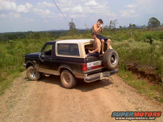 1990 Ford Bronco with two people sitting in the open rear, parked on a dirt road