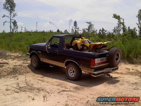Dark blue Ford Bronco parked on dirt road, yellow ATV in the bed. Trees and brush in background