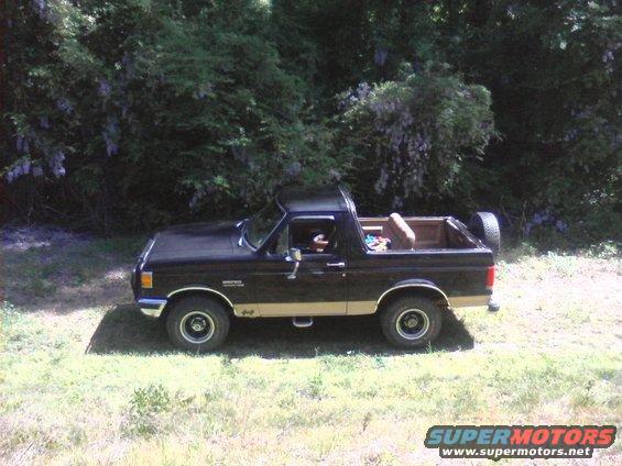 Side view of a black and tan 1990 Ford Bronco parked on grass with trees behind it