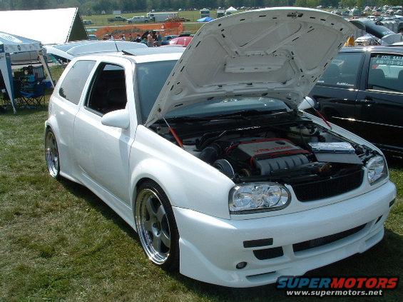 White hatchback with hood open, custom body kit, aftermarket wheels, and visible engine