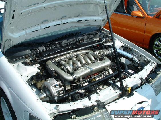 White car's engine bay with V6 engine and turbocharger. Orange car visible in background