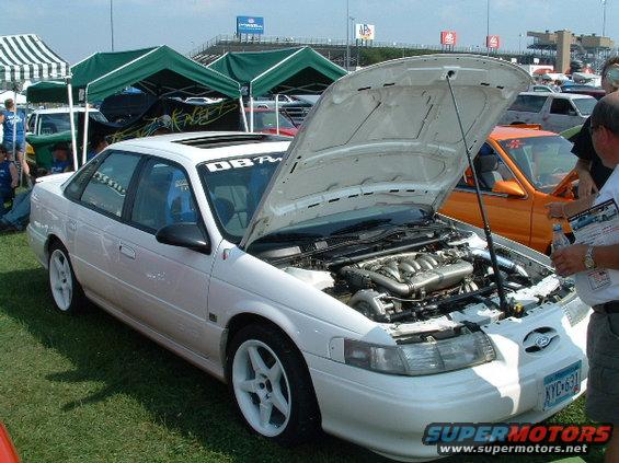 White sedan with hood open, showing engine and turbo. Blue racing seats visible inside. Other cars and tents in background