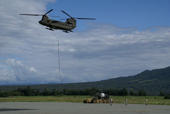 Military helicopter in flight, suspended load below, ground crew visible near cargo