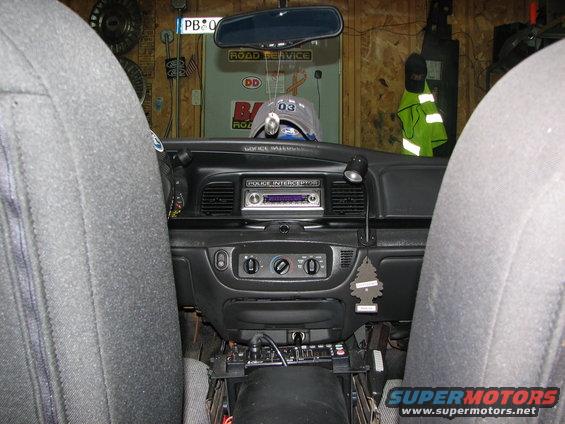 Interior view of a police car dashboard with a radio and air freshener. A hat with '03' is on the dash