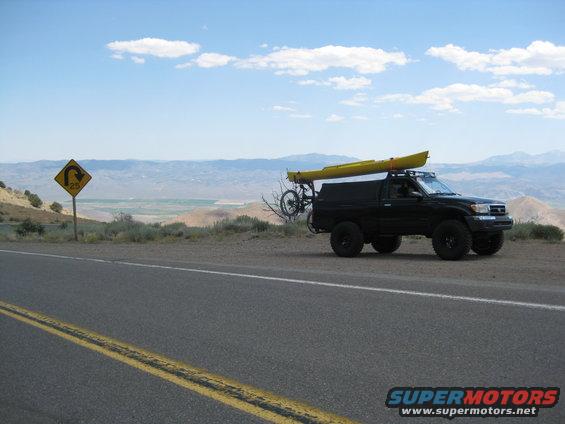 Black pickup truck with kayak on roof rack and bicycle on rear rack, parked on roadside with mountainous landscape in background