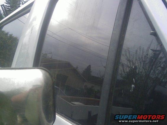Side view mirror and tinted window of a vehicle, reflecting houses and trees